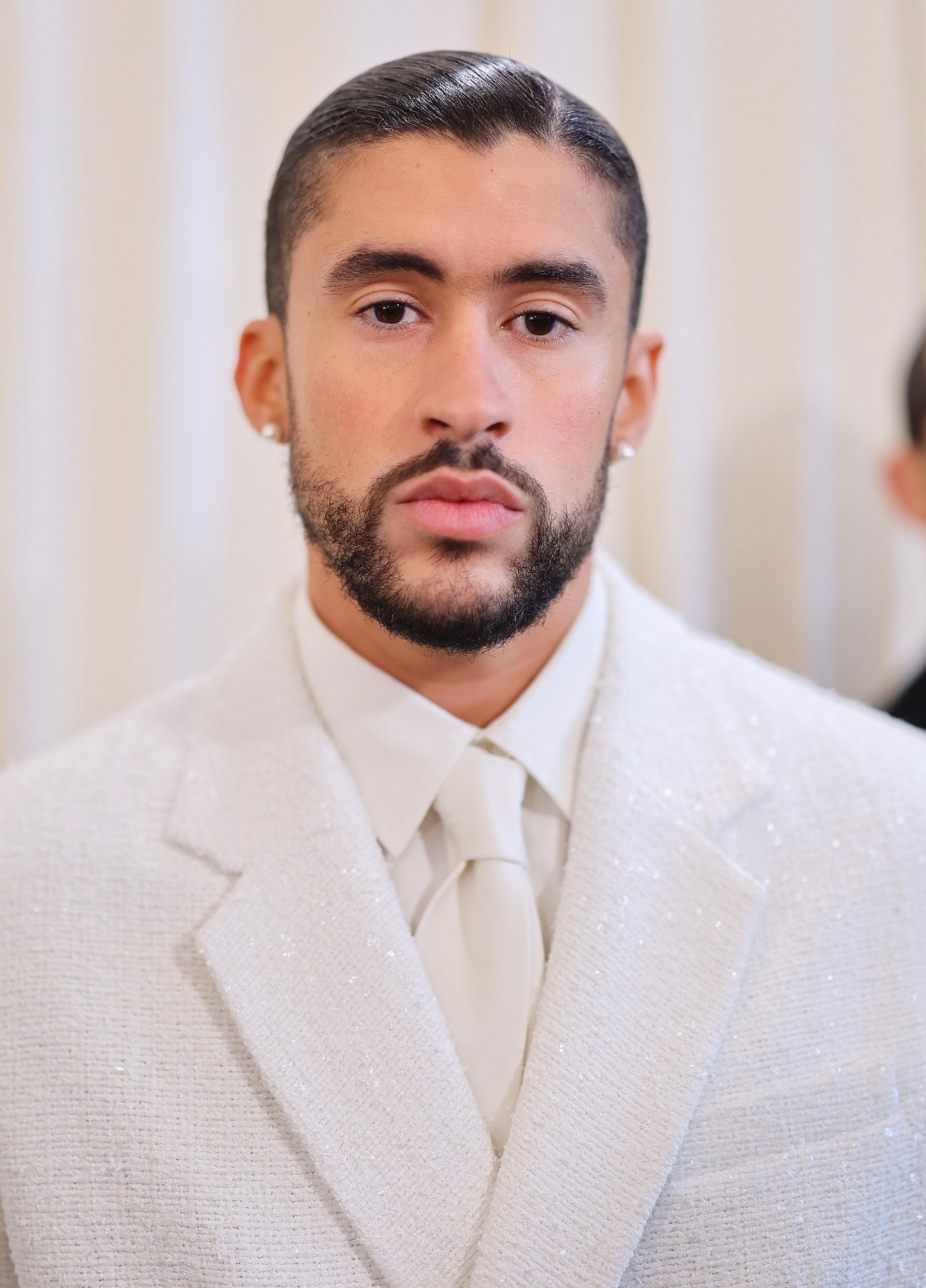 A person wearing a white, textured suit with a matching tie and earrings stands against a light-colored background.